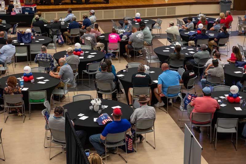 People seated at round tables in a large hall; some tables have decorations.