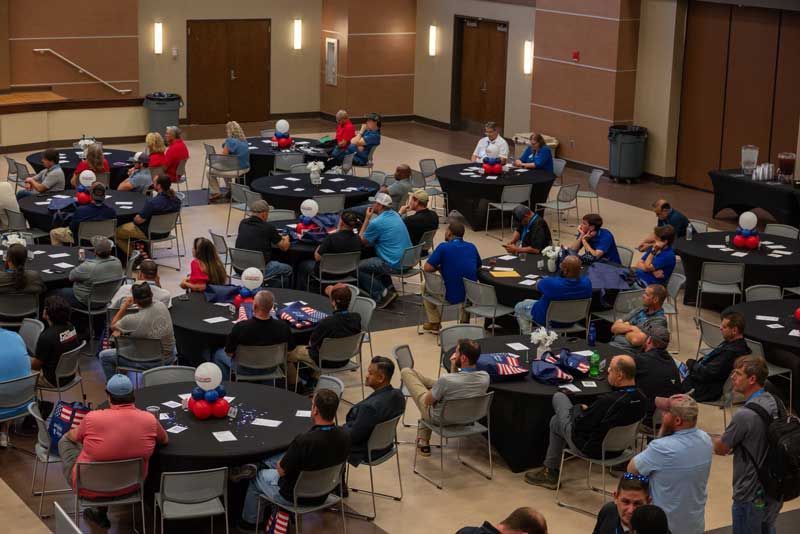 Large room with round tables, many people seated, mostly wearing blue.