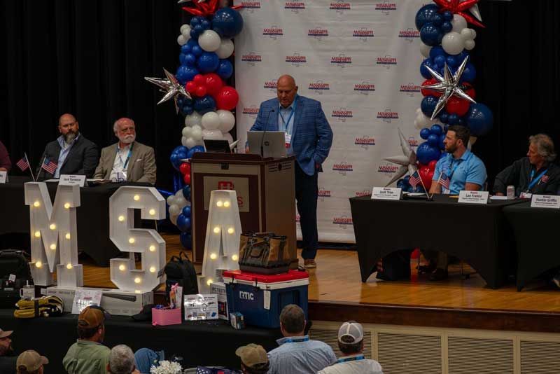 A man speaking at a podium on a stage with a panel of seated people.