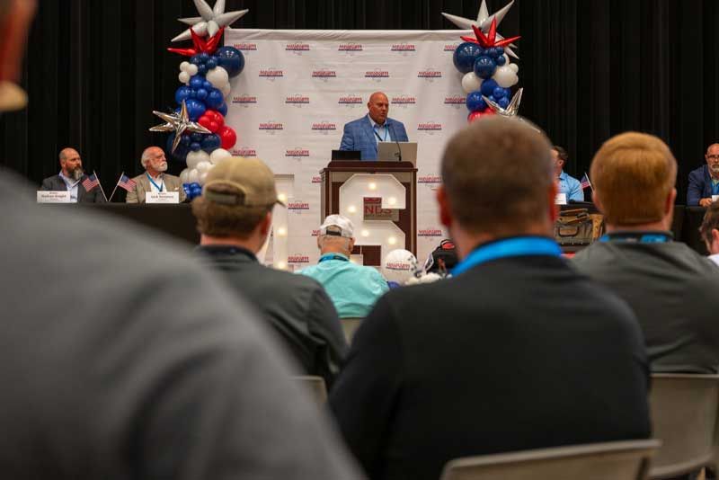 Man speaking at a podium, large event with audience, red, white, and blue decorations.