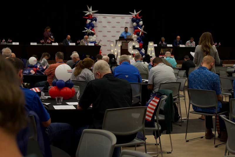 Political event with speaker at podium, crowd seated at tables, red, white, and blue decor.