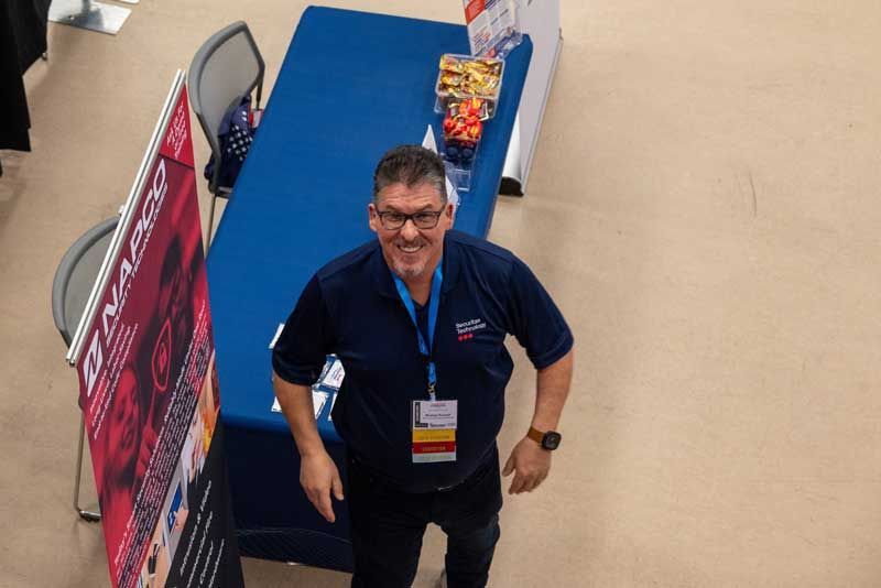 Man standing at a booth with a blue table. He wears a navy shirt and jeans, smiling. 