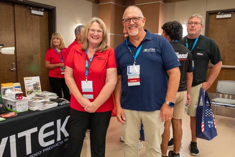 Two people stand in front of a table with products at a trade show. A woman in red and a man in blue smile.