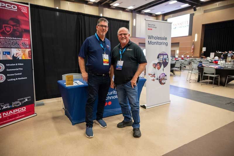 Two men stand at a trade show booth with banners; one blue, one white. They smile, both wearing lanyards.