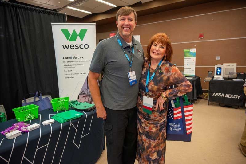 Man and woman pose by a Wesco banner at a conference. 