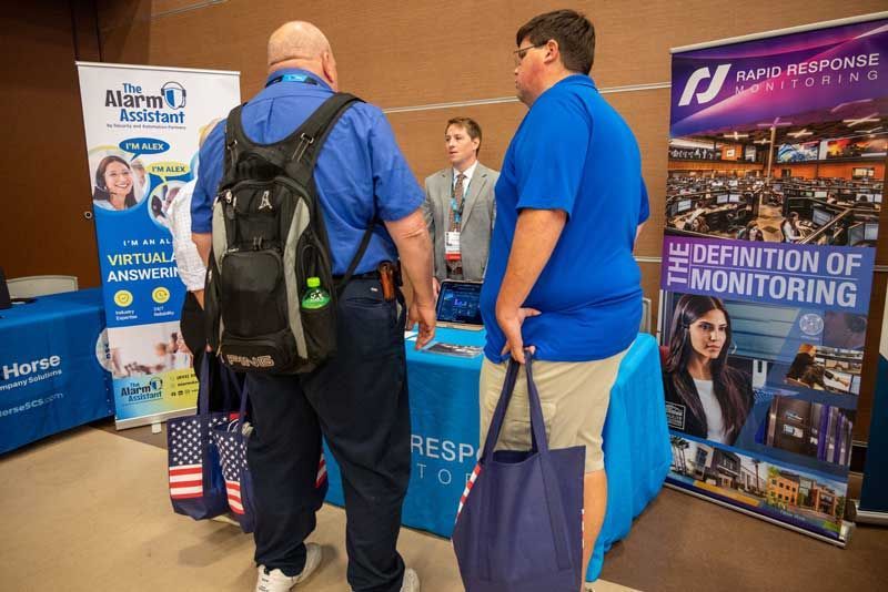 Two men in blue shirts at a trade show booth, looking at a representative behind a table; banners on either side.
