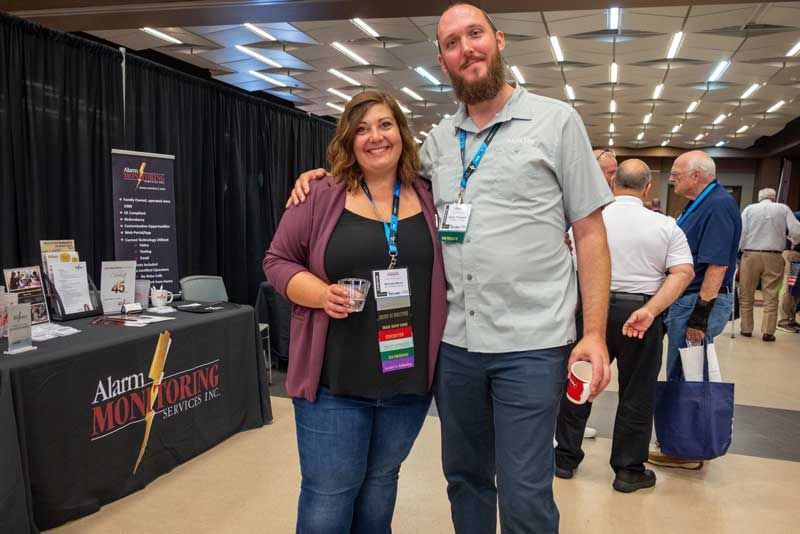 Woman with arm around a bearded man smiling at an event with a booth in the background.