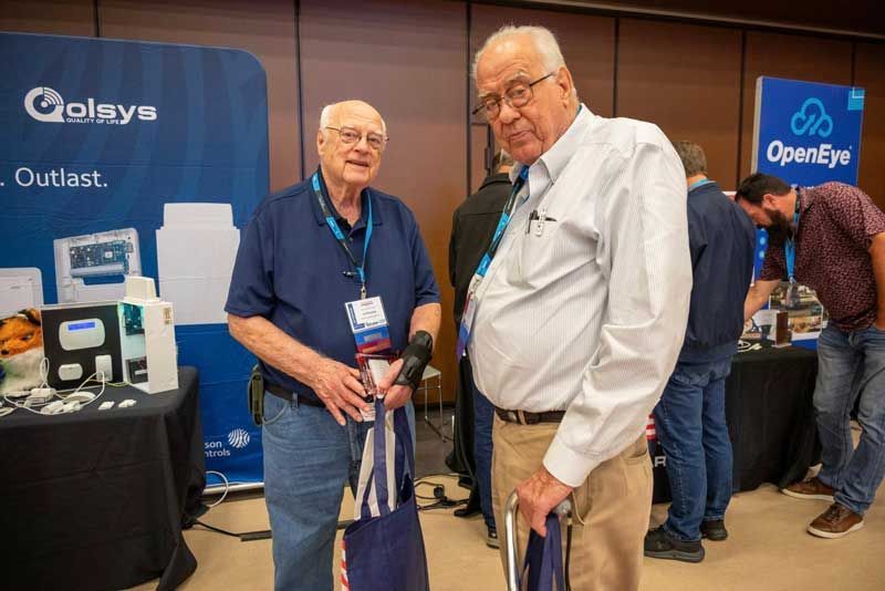 Two men at a trade show booth, one holding a bag, the other a cane. 