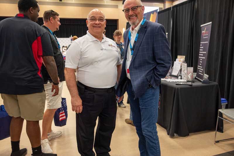 Two men smiling, standing at a booth with a table. One man in a polo shirt, the other in a suit jacket.