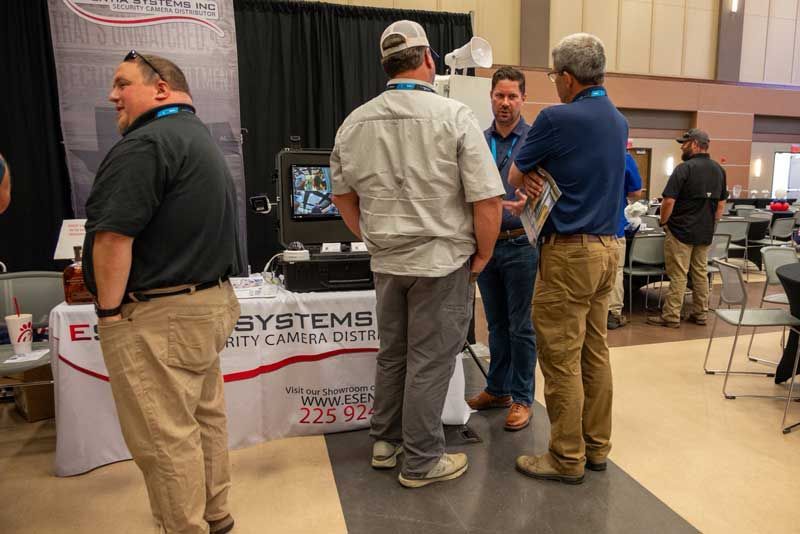 People at a trade show booth, viewing a security camera display. Men in casual clothes converse around the booth.