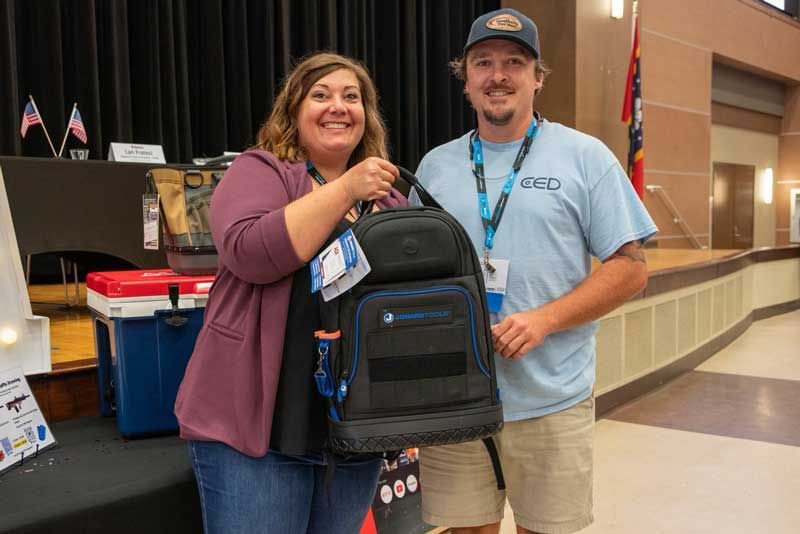 Woman and man hold a black and blue bag in an auditorium. Man wears a hat and lanyard. Woman smiles.