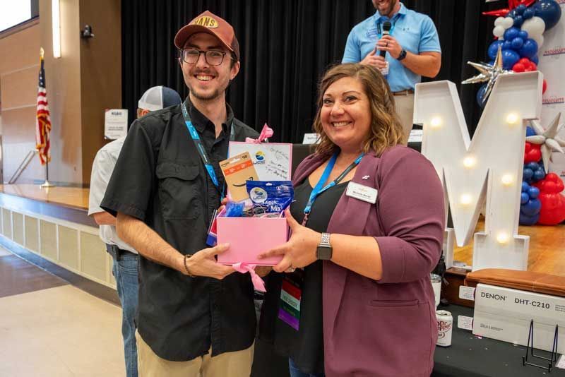 Two people smiling, holding a pink gift box; stage backdrop.
