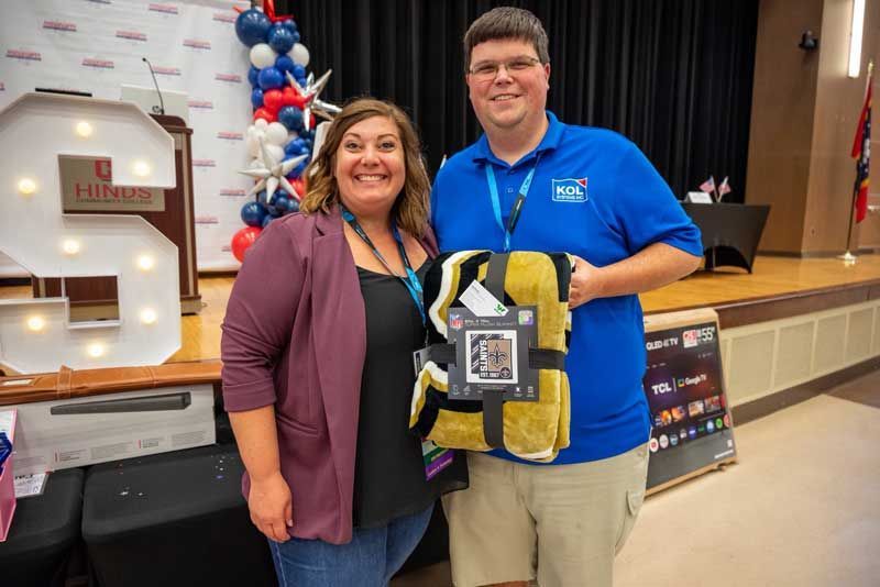 Woman and man holding a blanket with a label, indoors on a stage. Balloons and a lit 
