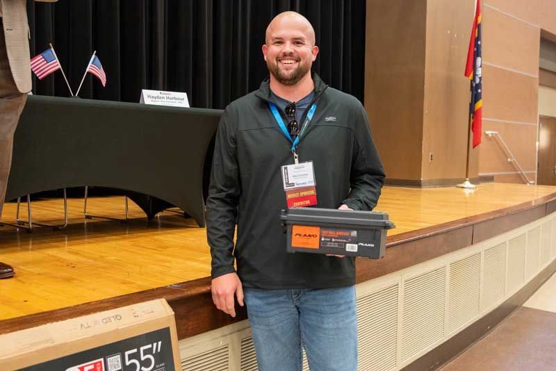 Man smiles, holding a box, standing on a stage with a table, flags, and a TV box.