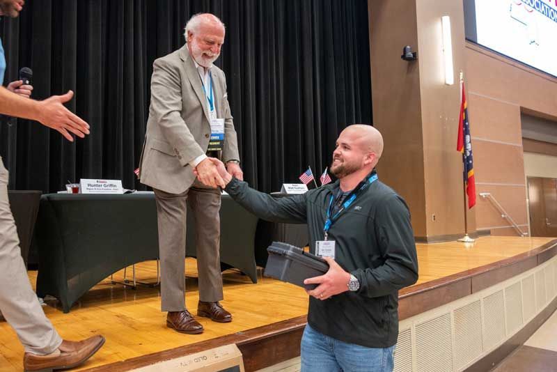 Man in blazer shakes hands with another man, accepting a box on a stage with a table and US flags.