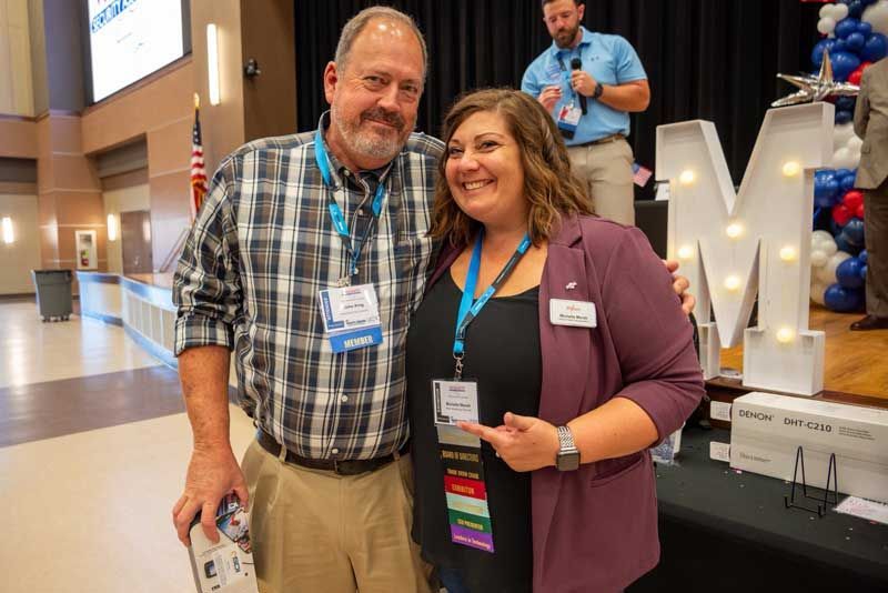Man and woman smiling, posing for a photo. They are in a conference hall with an illumin