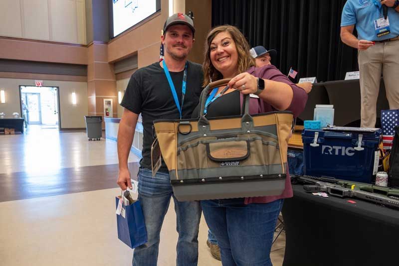 Two people smile, one holding a gift bag, the other a large tote bag, at an event with tables and signs.