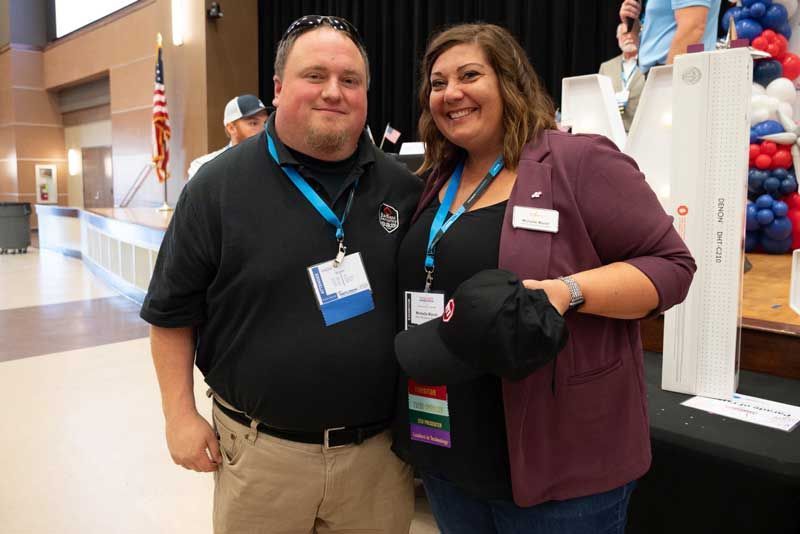 Two people smiling together at an event. Man in black shirt, woman in burgundy blazer. American flag visible in background.