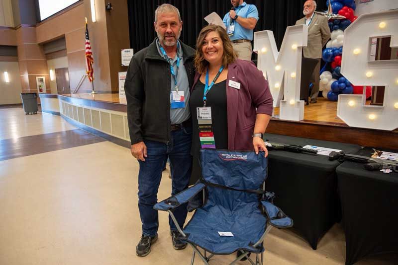 Two people holding a blue folding chair, standing in a large room with a stage.