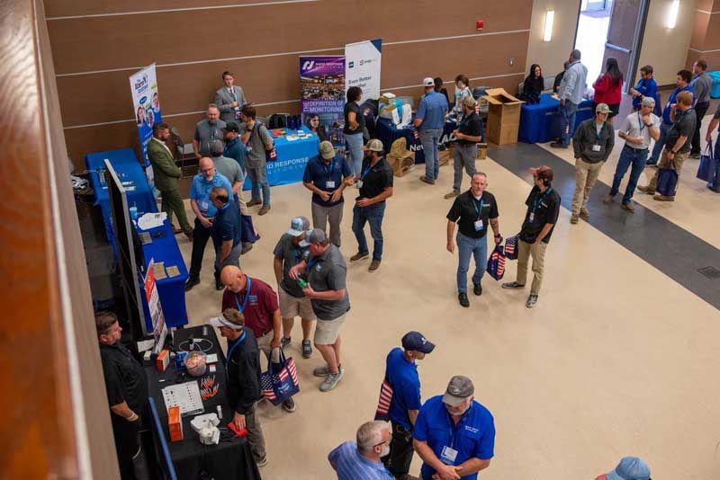 People at a conference, some at booths with displays, in a large room with blue and brown accents.