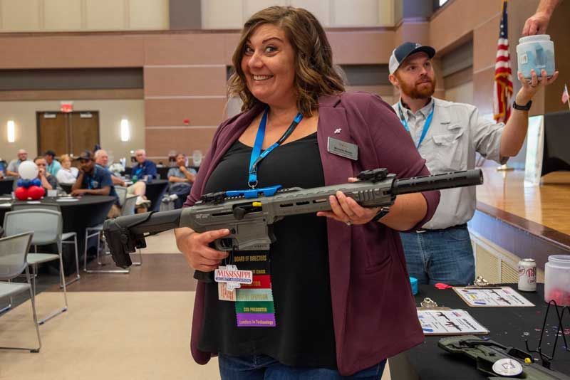 Woman smiles holding a gray rifle at an indoor event.