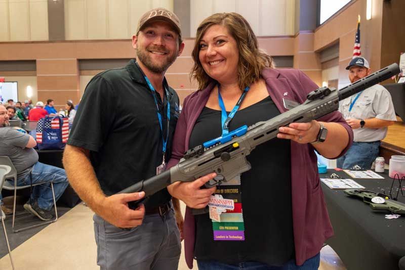 Man and woman smiling, holding a gray firearm in a convention hall, with attendees visible.