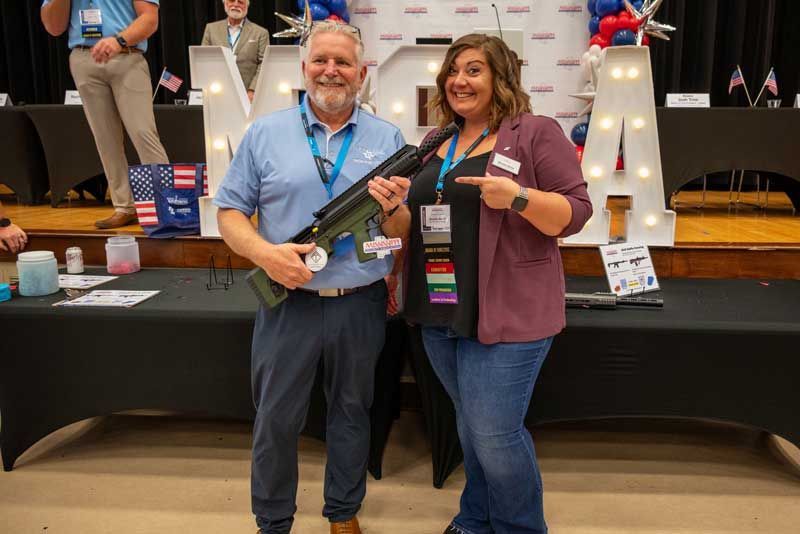 Man and woman holding a green firearm at a table with political decorations; woman points at the weapon.