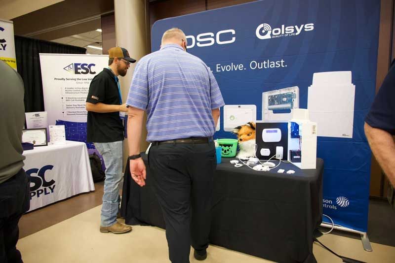 People at a trade show booth with security system displays. A person examines the merchandise. Blue backdrop.