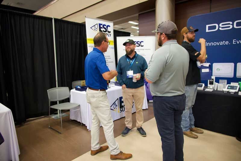Four men converse at a trade show booth, likely discussing security products.