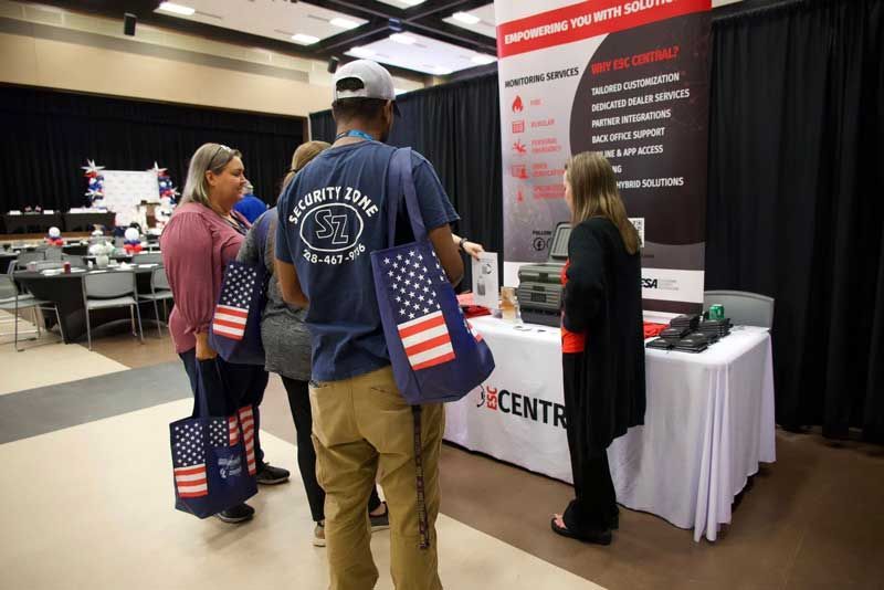 People at a vendor table with a banner that lists security services; a few people hold American flag-themed tote bags.