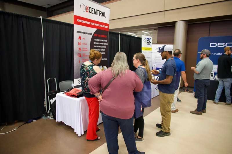 People at a trade show booth with a banner. A table and black backdrop. Several people are engaged in conversation.