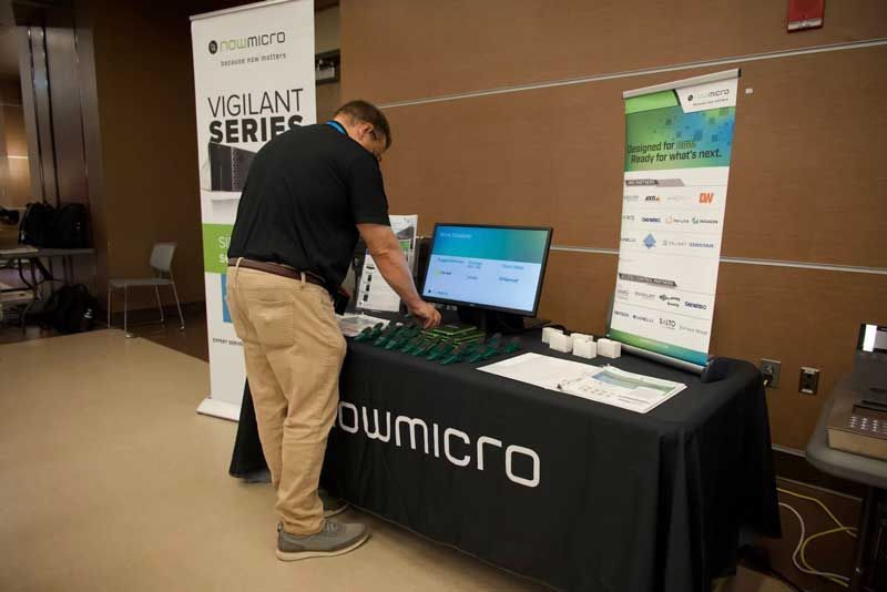 Man at a conference booth, arranging items on a table with a computer monitor and promotional banners.