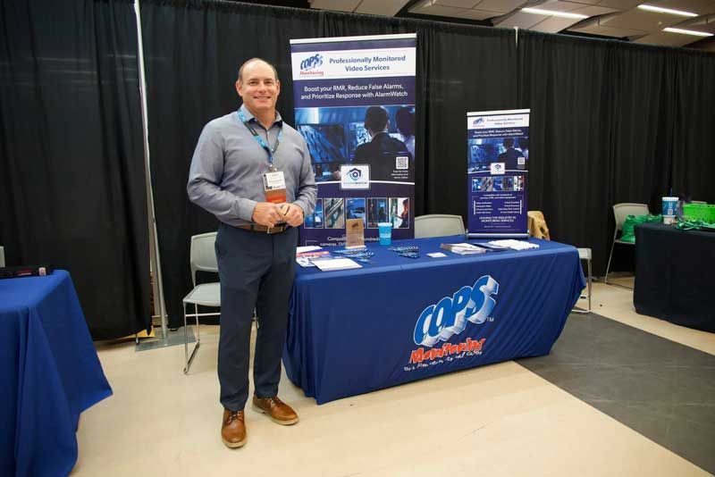 Man stands at a trade show booth, smiling. Booth has a blue table cover and banner.