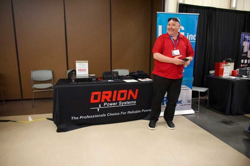 Man in red shirt stands at Orion Power Systems display, smiling. Brown and black background at a trade show.