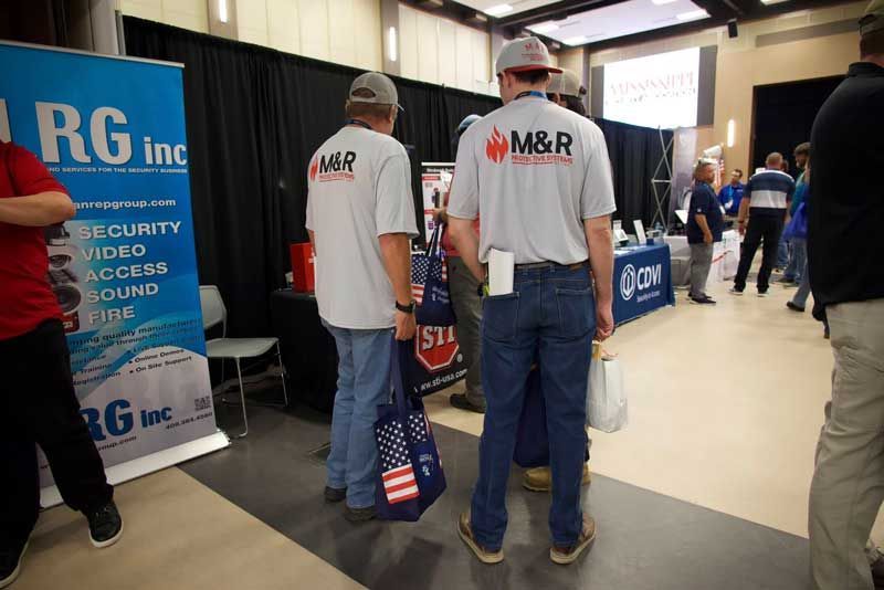 Two men wearing M&R shirts at a trade show. They're facing away, near a booth with security info.