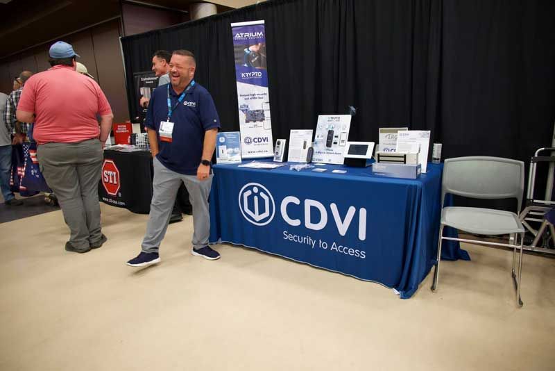 Man at a CDVI security booth at a trade show. Products displayed on table. Others stand nearby.