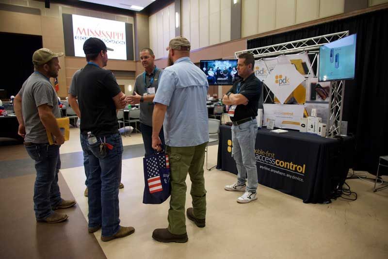 Men at trade show booth discussing products, screens displaying data, black table with logo.