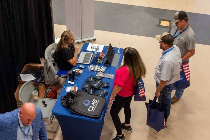 People at a booth with blue table. Woman speaks to attendees, CMS logo on bags.