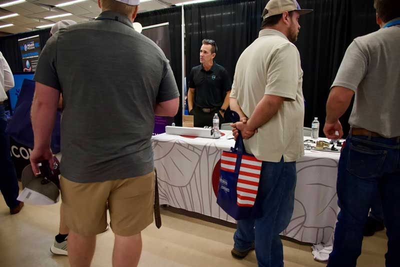 People at a booth with a vendor. One person holds a patriotic bag. White table with items.