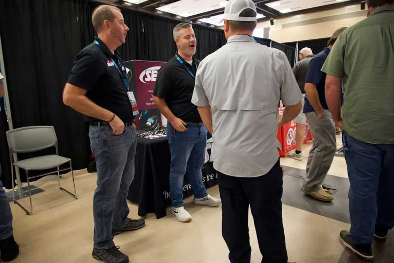 Three men at a booth, talking. One in a gray shirt, hat, facing away. Others wear black shirts, jeans.