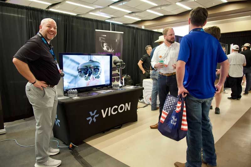 Man smiling by Vicon display at a trade show; people converse nearby.