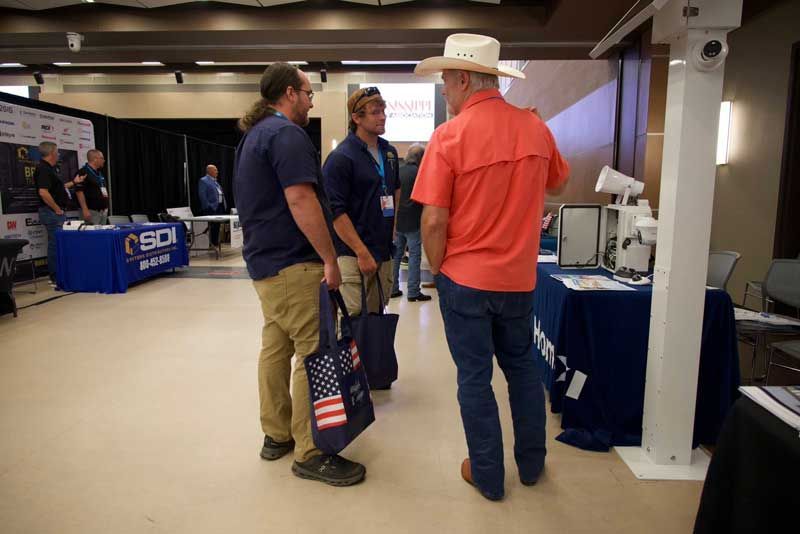 Three men in casual attire converse at an event, with vendor booths in background.