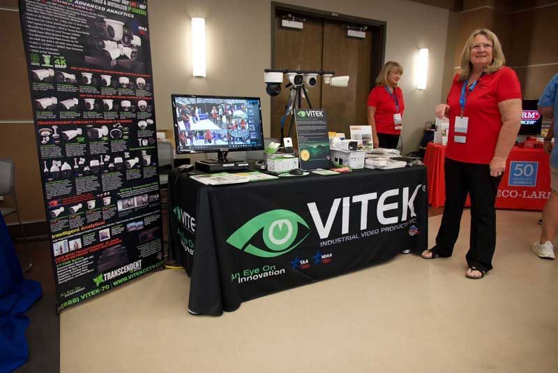 Vitek security booth with monitor, cameras, banner, two women in red shirts, conference setting.