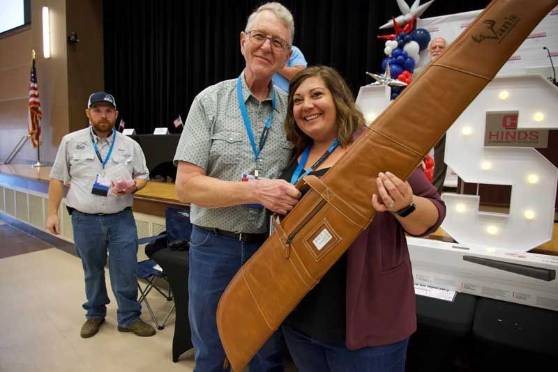 Man and woman smiling, holding a long, brown rifle case indoors. 