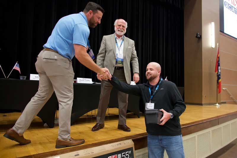 Man in blue shirt shakes hands with man holding a box on a stage. 