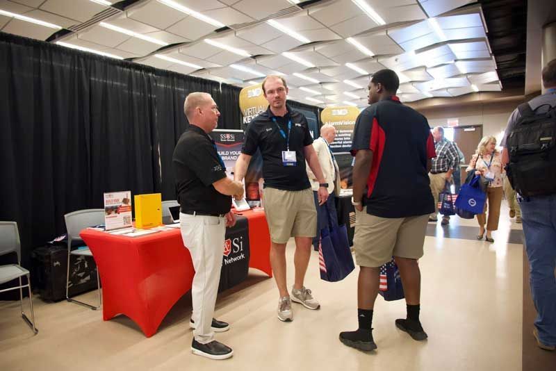 Three men talking at a trade show booth, a man on left shakes hands, other two stand nearby. 