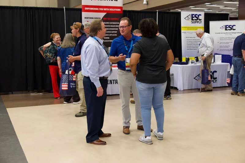 People in conversation at a trade show booth; two men and a woman talk, others stand nearby.