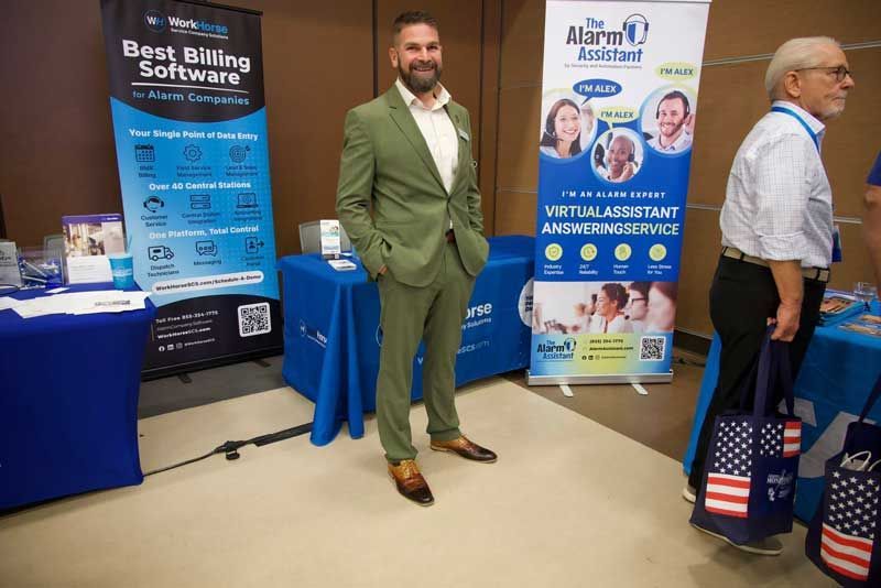 Man in green suit stands at a business expo booth, near blue banners and a table.