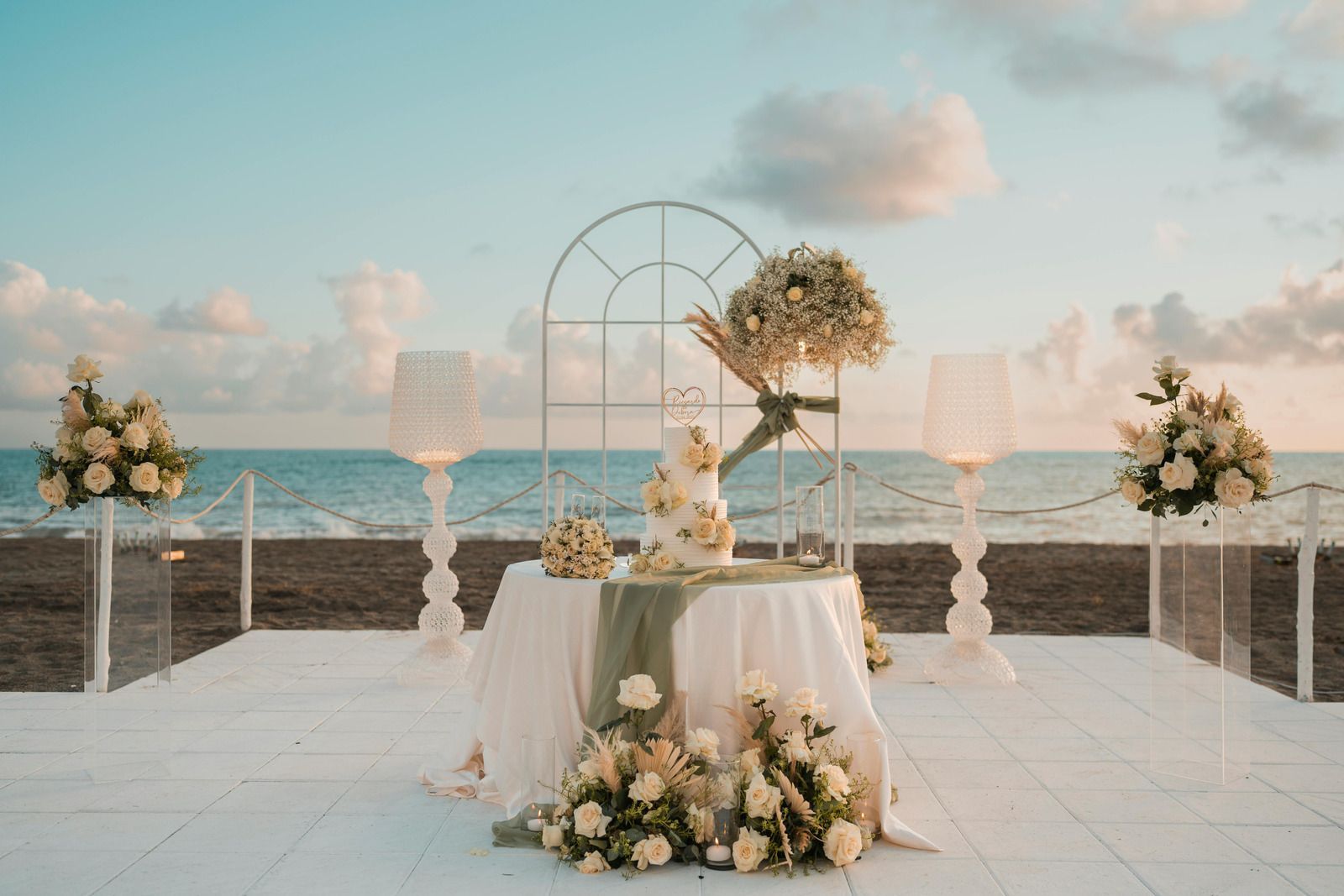 Beachfront wedding ceremony setup with white floral arch, table, and aisle overlooking the sea