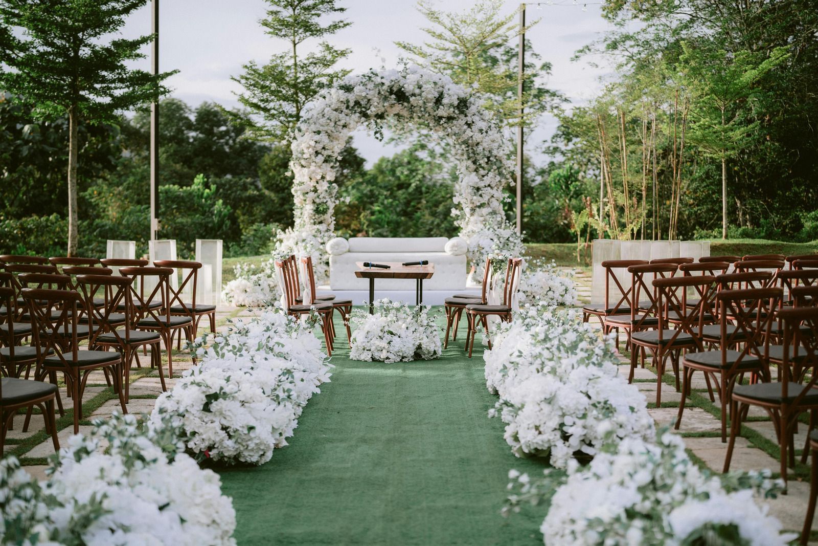 Outdoor wedding ceremony setup with white floral aisle, green runner, and wooden chairs facing an altar.